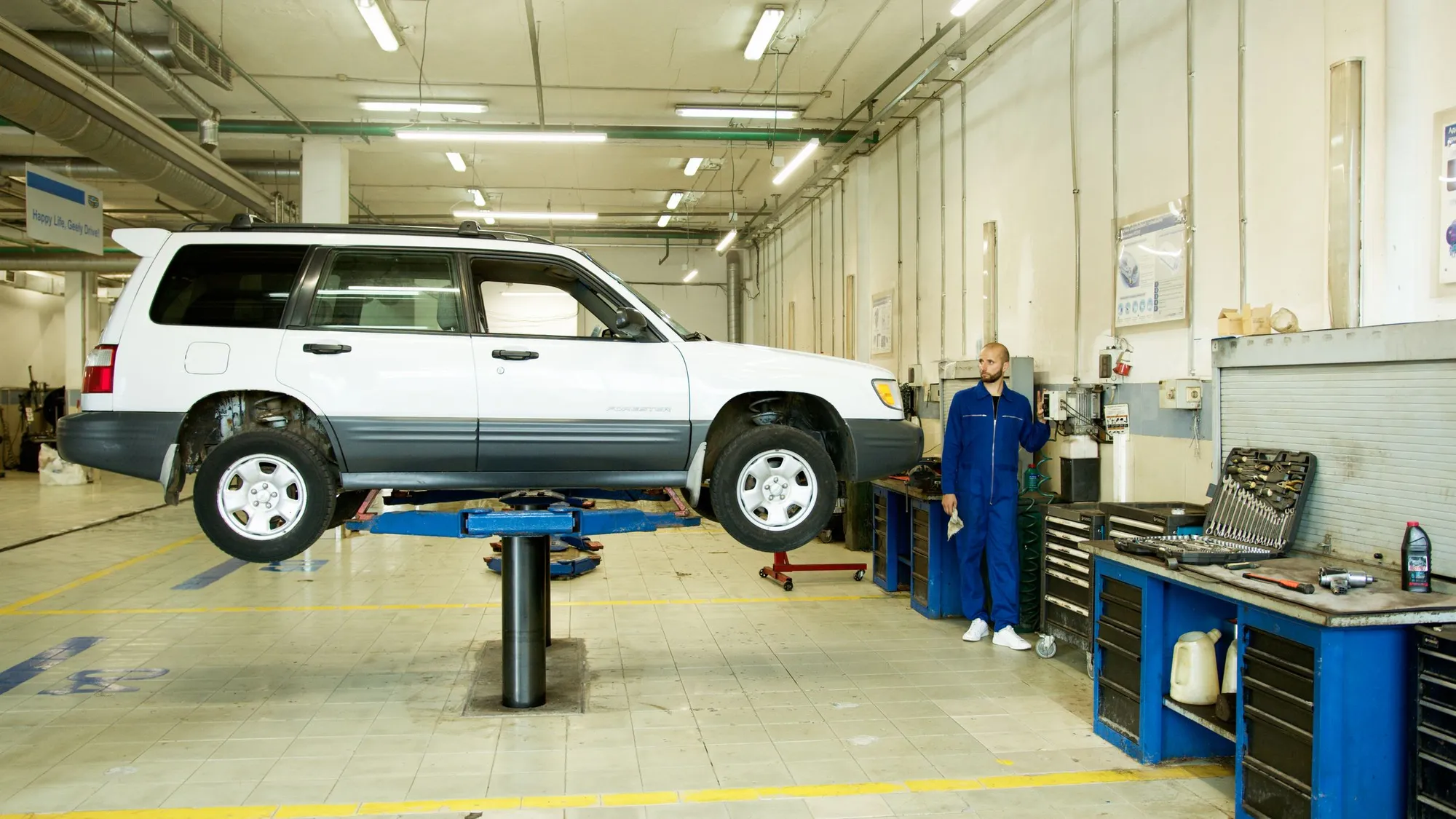 Nave del taller con un todoterreno blanco subido al elevador, un mecánico al fondo y los bancos de herramientas azules